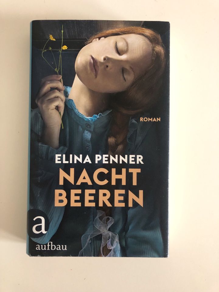 Couverture du roman Nachtbeeren d'Elina Penner. Une femme en robe bleue, avec une tresse rousse, les yeux clos, le visage penché, avec le cou penché à 90 degrés, tient un brin de boutons d'or dans une main tenue en l'air.