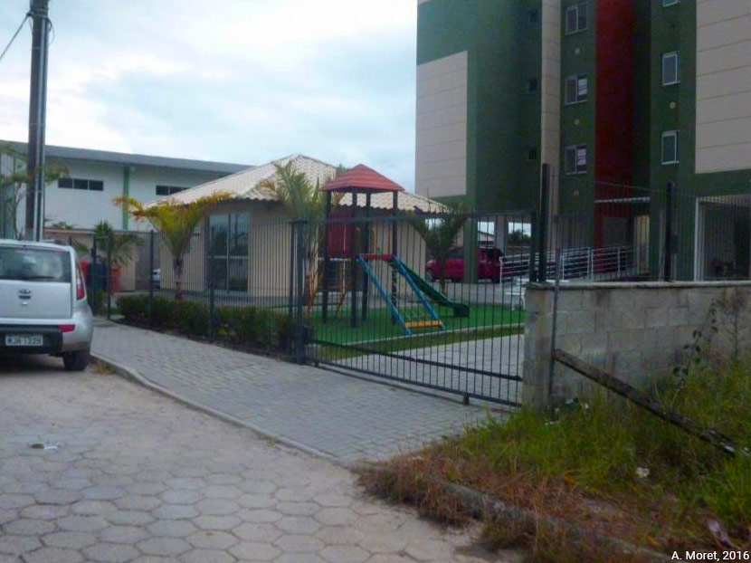 Vue de la résidence Portal do Sol, depuis la rue. La résidence correspond à deux immeubles verts et blancs. Les jeux pour enfants visibles sur la pelouse au centre de la photo sont très utilisés. Photographie prise à Ponta de Baixo, Biguaçu, en mars 2016 par Alice Moret.