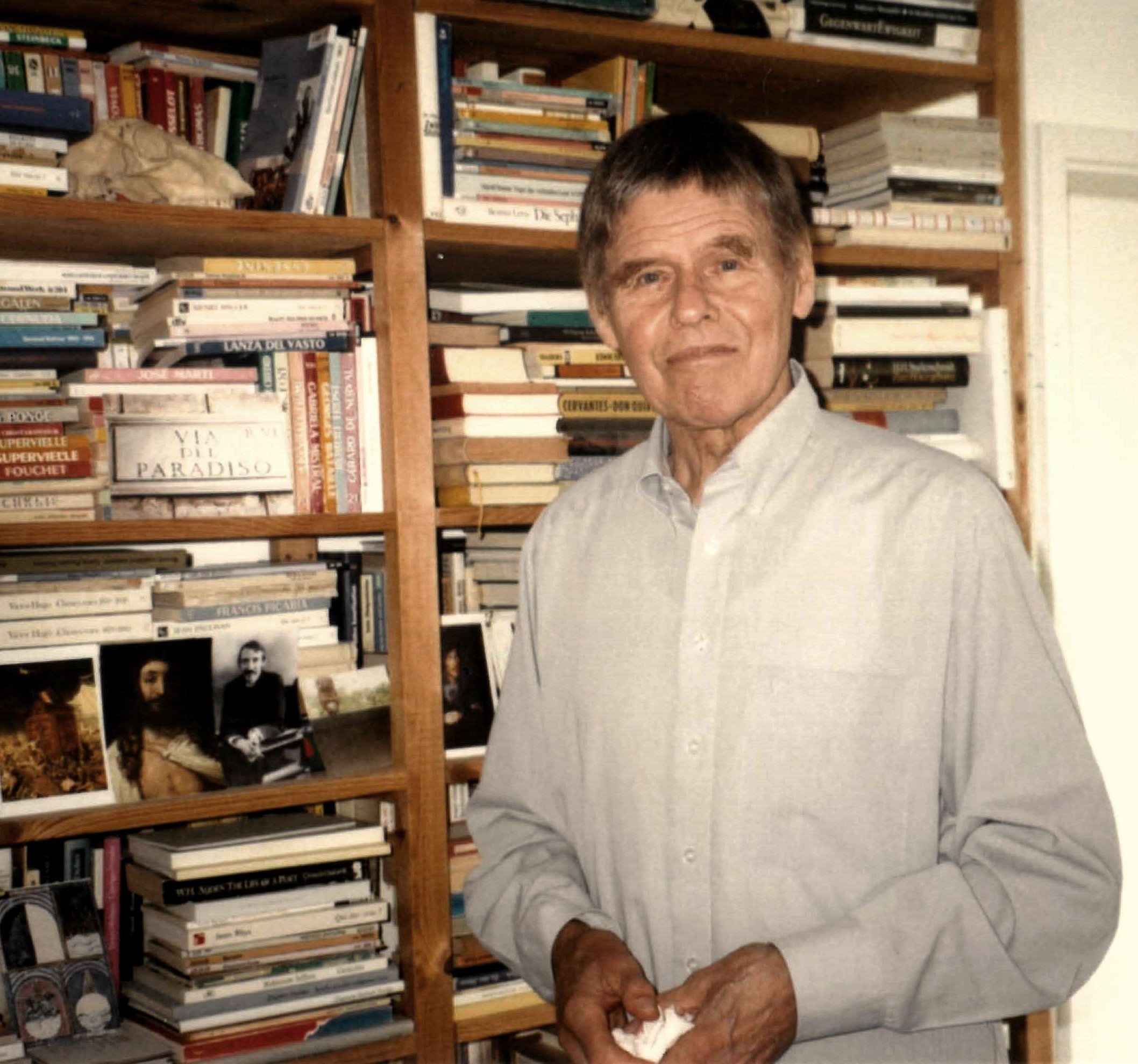 Portrait de l'auteur Christoph Meckel. Souriant, celui-ci se tient, au premier plan de l'image, devant une bibliothèque aux rayons surchargés de livres et agrémentés de quelques photographies. 
